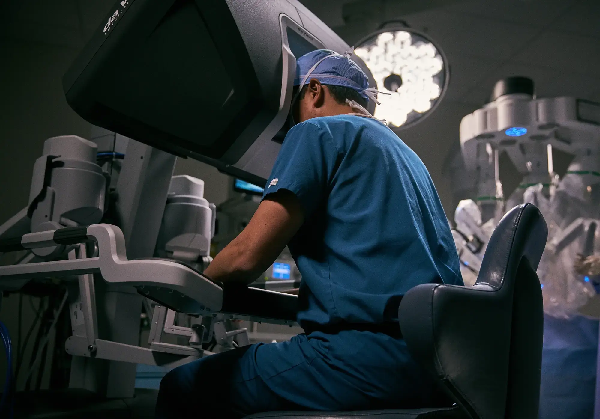 Dr. Wei Phin Tan performing robotic prostatectomy at the da Vinci surgical console at NYU Langone, blue scrubs and surgical cap, operating room lights and robotic arms visible